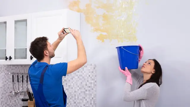 Homeowner photographing a stained wall while a second person holds a bucket under a leak