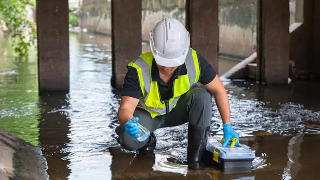 Worker in a hard hat and gloves taking a water sample from dirty standing water